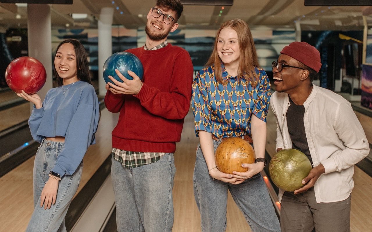 Symbolbild Bowling: Vier Freunde lachen mit bunten Bowlingkugeln auf der Bahn