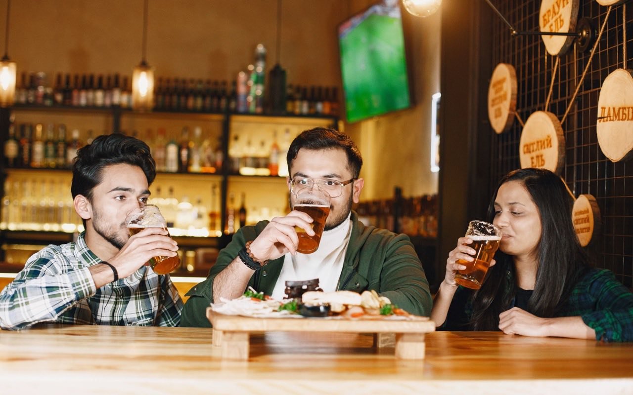 Symbolbild Brauereiführung/Bierverkostung: Drei Erwachsene probieren Craft Beer in Bar mit Holzeinrichtung